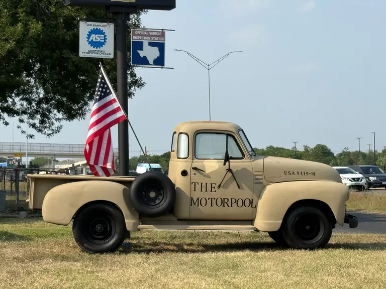Side view of the vintage tan Motorpool truck beside the ASE Certified Professionals and Texas official vehicle inspection station signs, with an American flag